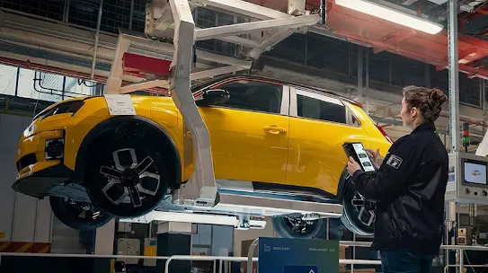 A worker using a tablet in front of a yellow car suspended on an assembly line.