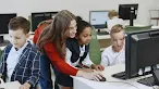 A teacher in a red sweater leans over a desk to assist two young students working together at a desktop computer in a classroom.
