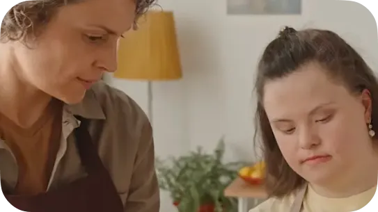 A woman with curly hair watching a young woman with Down syndrome attentively while working together indoors.