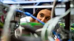 A woman looks intently through a complex array of wires, cables, and structural frames of a piece of technical machinery or computing equipment.