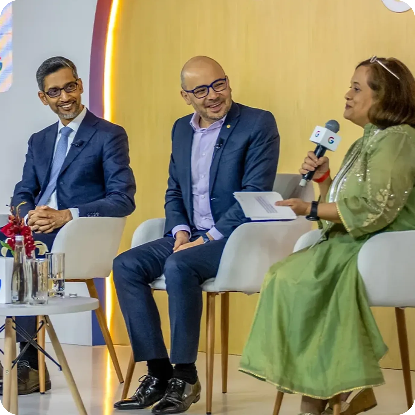 Sundar Pichai and two other executives sitting in white chairs on a stage for a panel discussion. A woman on the right holds a Google-branded microphone and addresses the group.