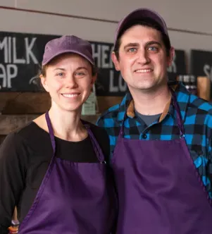 A smiling man and woman wearing matching purple baseball caps and aprons, standing in front of a chalkboard sign in their New Hampshire soap shop.