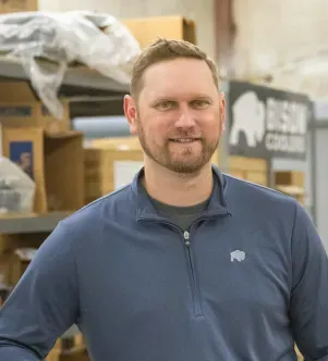 A smiling man with a goatee wearing a blue quarter-zip pullover with a small bison logo, standing in a warehouse with a Bison Coolers sign and shelves of boxes in the background.