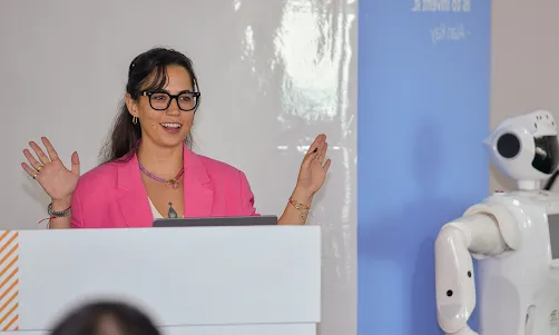 The female presenter, dressed in a pink suit, is leading the event and delivering her segment.