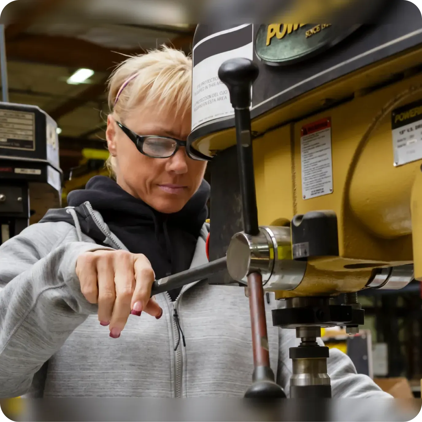 A blonde woman wearing safety glasses and a grey hoodie operating a yellow Powermatic industrial drill press in a manufacturing workshop.