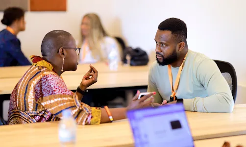 A women holding mobile on hand and discussing something with a man who is seated in front of her.