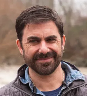 Close-up portrait of a man with a dark beard and hair, smiling outdoors with a blurred river and trees in the background, featured in a Maine Fly Company growth story.