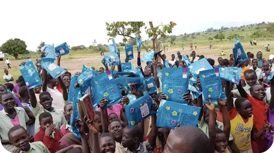 A large crowd of young students outdoors cheering and holding blue "Days for Girls" reusable sanitary pad kit packages in the air.