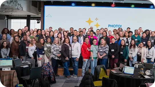 A large, diverse group of educators and professionals posing for a photo at the AI Literacy Day event. They are standing in front of a large screen displaying yellow stars and the word "PHOTO!". Laptops and round tables are visible in the foreground.