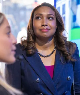 A woman wearing a blue jacket facing away from the camera and smiling and another woman in soft focus and discussing with a man.