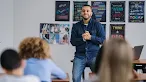 A smiling male teacher stands at the front of a classroom, clasping his hands together as he engages with the students seated before him.