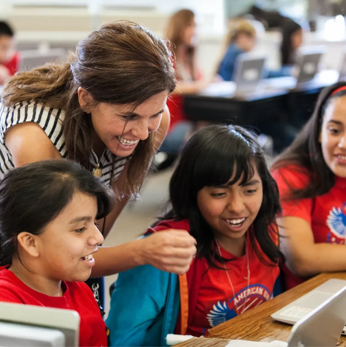 A smiling female teacher leans over and points at a laptop screen, engaging with three young female students who are also smiling in a classroom.