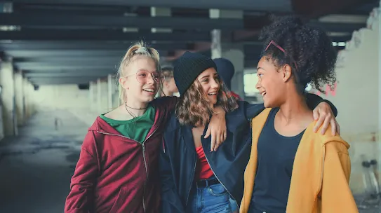 Three smiling teenage friends walking with their arms around each other inside an urban warehouse-style building.