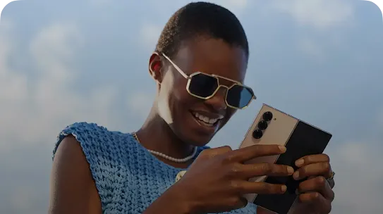 A smiling person with short dark hair, wearing geometric sunglasses, a pearl necklace, and a blue knit sleeveless top, holding a partially folded smartphone horizontally against a partly cloudy sky.