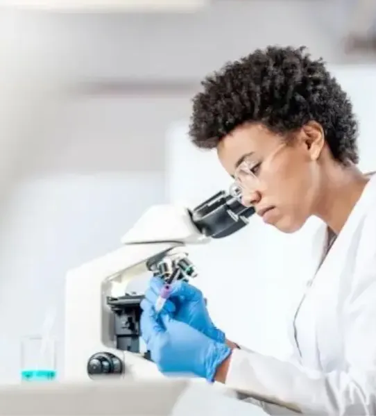 A female scientist in a white lab coat and blue gloves focuses on a sample vial while using a microscope in a bright, modern laboratory setting.