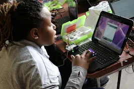 A close-up shot of a student with braided hair typing on a laptop, which is connected to a circuit board and various electronic sensors from a Thimble.io kit.