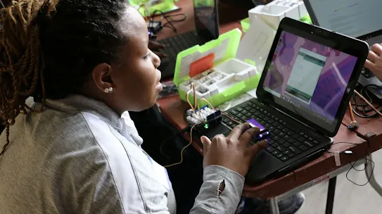 A close-up shot of a student with braided hair typing on a laptop, which is connected to a circuit board and various electronic sensors from a Thimble.io kit.