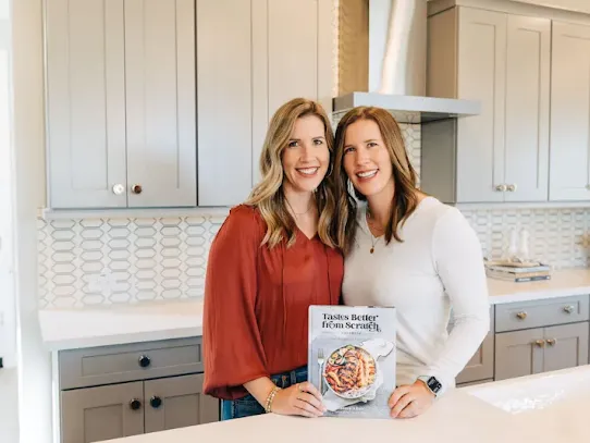 Lauren and Liz in their kitchen holding their cookbook.