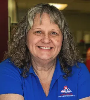 A smiling woman with curly graying hair wearing a royal blue polo shirt featuring the Custard Stand Chili logo in an indoor setting.