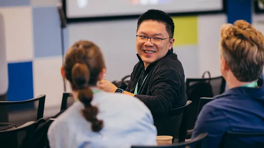 A smiling man with glasses chatting with colleagues during a presentation or conference session.
