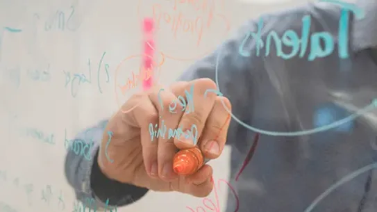 A close-up shot of a person’s hand using an orange marker to write brainstormed words like "Partnership" and "Talents" on a clear glass whiteboard. The background is blurred, showing a modern office environment.