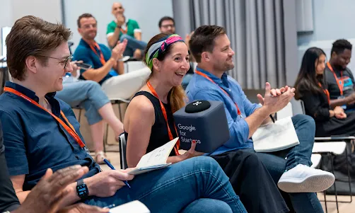 Un grupo de asistentes sentados en un taller, sonriendo y aplaudiendo, con una persona sosteniendo un dispositivo con la marca Google for Startups.