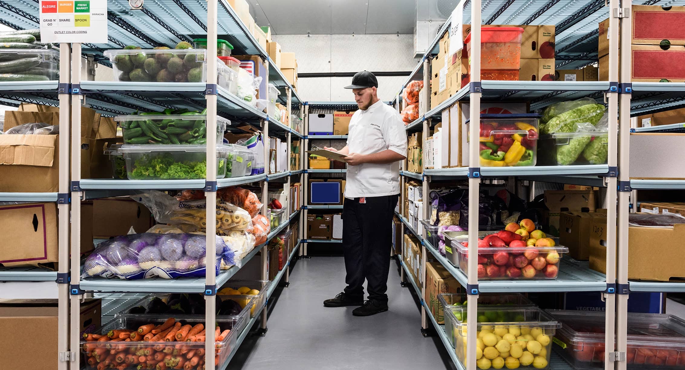A man with a clipboard examines stacks of vegetables and fruit.
