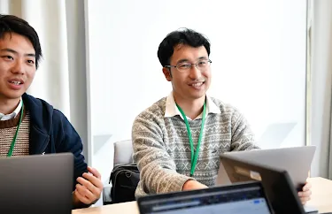 Two individuals wearing ID badges around their necks, seated in a conference room listening to a speaker.