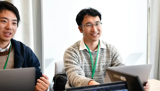 Two individuals wearing ID badges around their necks, seated in a conference room listening to a speaker.