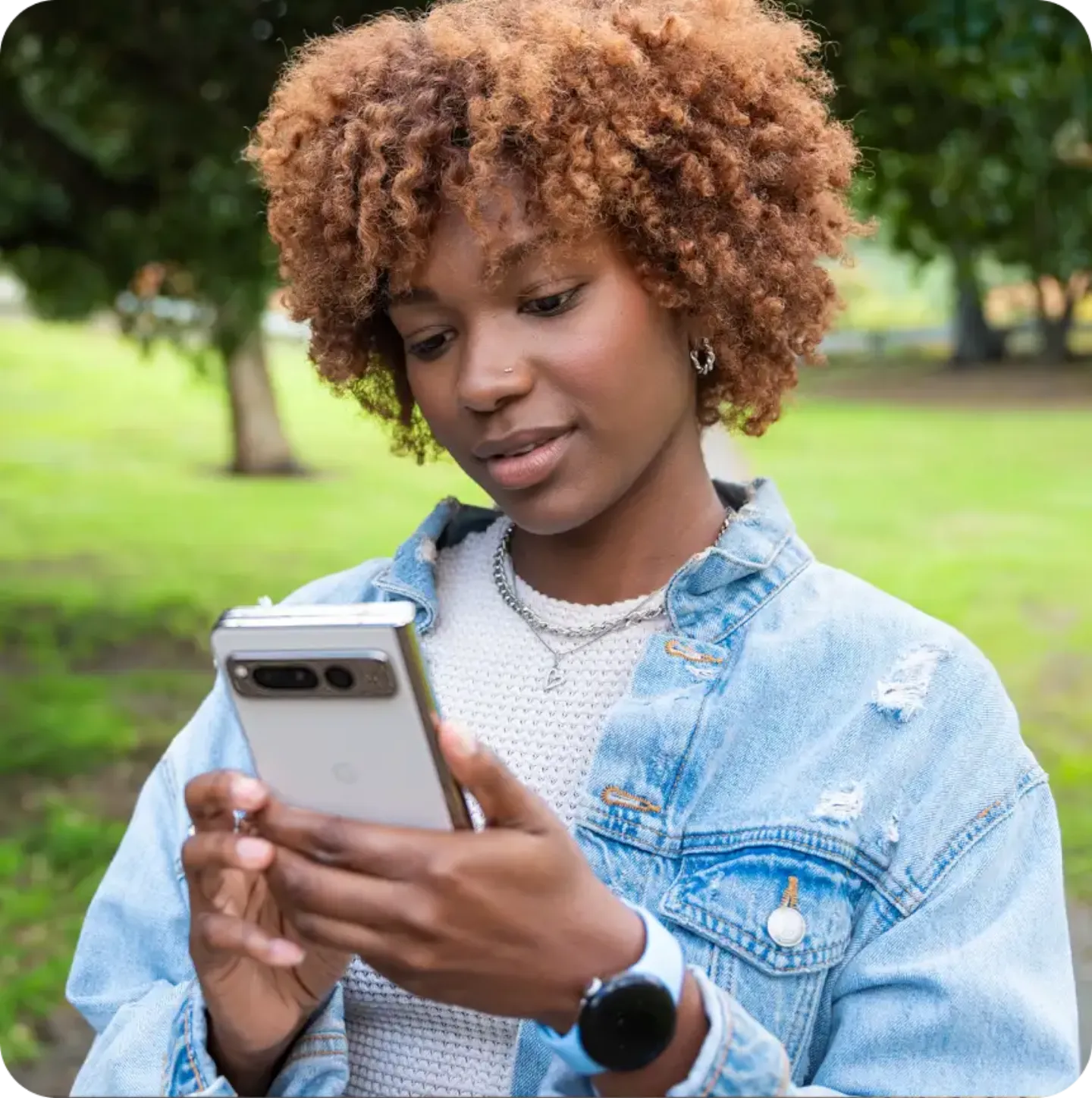 A young woman with curly, reddish-brown hair looks down at her smartphone while standing outdoors in a park.