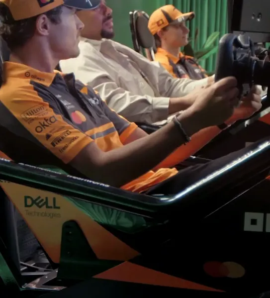 Lando Norris and Oscar Piastri are talking and gesturing to another person in a light-colored shirt. They are having a friendly, competitive conversation about getting practice laps on the simulator. The background features a green curtain.