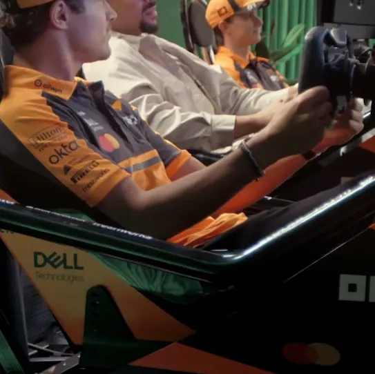 Lando Norris and Oscar Piastri are talking and gesturing to another person in a light-colored shirt. They are having a friendly, competitive conversation about getting practice laps on the simulator. The background features a green curtain.
