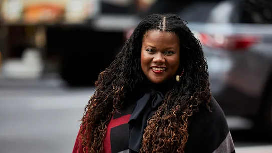 A professional portrait of Dara Byrne, Dean of Macaulay Honors College, smiling in a New York City setting. She has long, dark micro-braids with auburn highlights and is wearing a black pussy-bow blouse and a red, black, and grey patterned wrap. The urban background is softly blurred with a vehicle and city street visible.