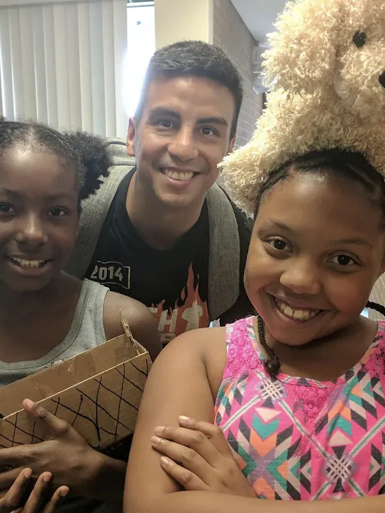 Thimble.io founder Oscar Pedroso smiles alongside two young students who are proudly showing off their science projects in a classroom setting.