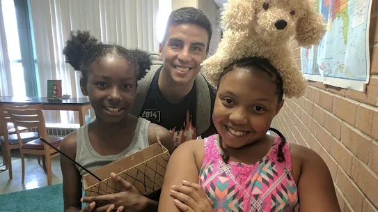 Thimble.io founder Oscar Pedroso smiles alongside two young students who are proudly showing off their science projects in a classroom setting.