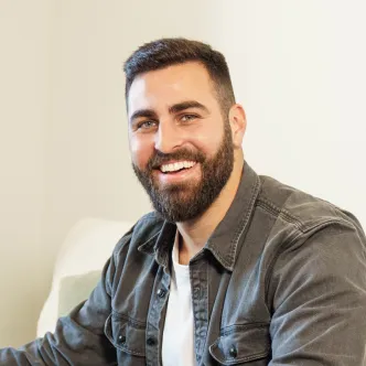 A man wearing khakis and a brown button down sits on a cream couch holding his laptop and looking down at his golden retriever