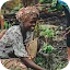 A smiling woman in vibrant patterned clothing and a headwrap kneeling in a field to pull weeds while carrying a sleeping baby on her back.