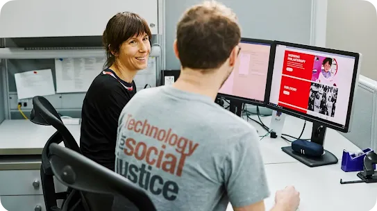 A woman in a dark shirt smiling at a male colleague whose gray t-shirt reads "Technology for Social Justice" as they review website designs on dual desktop screens.