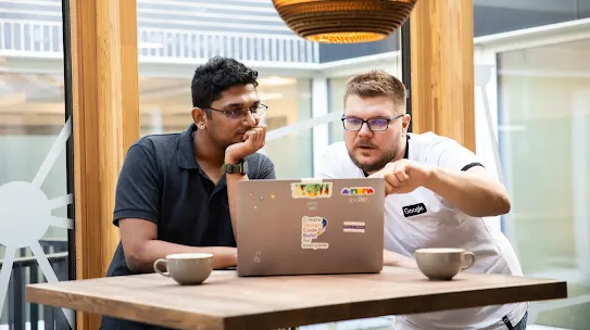 Two men are collaborating over a laptop at a high wooden table in a brightly lit, modern office space. On the left, a man in a dark polo shirt leans in with his hand on his chin, looking intently at the screen. Beside him, a man in a white "Google" t-shirt points toward the laptop screen while explaining something. Two cups of coffee sit on the table, and a large, textured pendant light hangs above them. The background features large windows showing an outdoor courtyard or walkway.