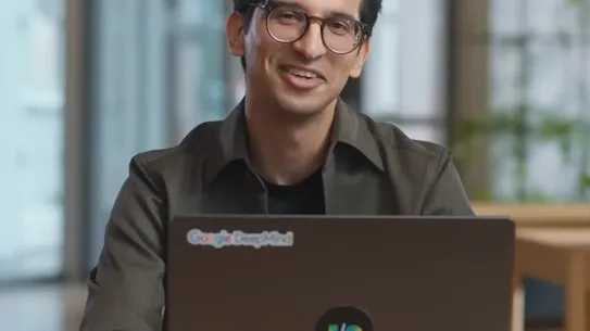 A person with glasses and dark hair is seated at a table and smiling, with a "Google DeepMind" logo visible on the top left of their laptop