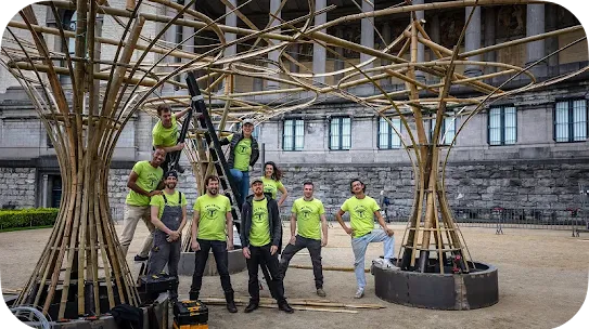 A group of eight construction or design team members wearing matching neon green t-shirts that say "THE TRIBE" pose together outside. They are standing around and climbing a ladder on a large, complex architectural pavilion made of curved bamboo poles. A historic stone building with columns stands in the background.