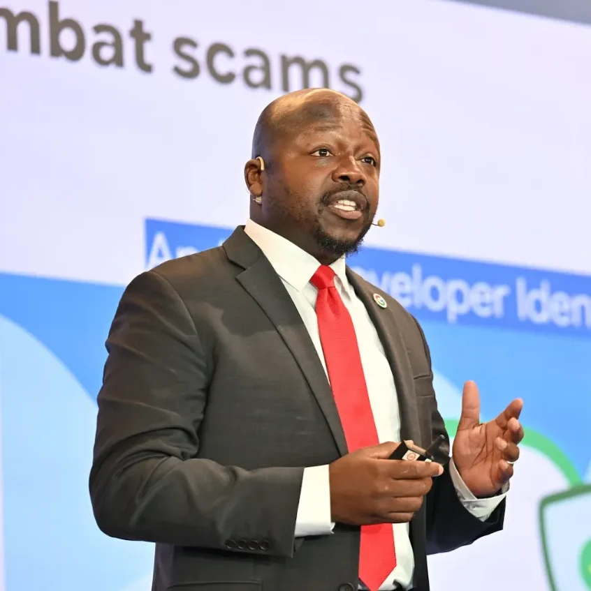 A man wearing a jacket and a red tie stands on stage speaking with the words "combat scams" displayed behind him.