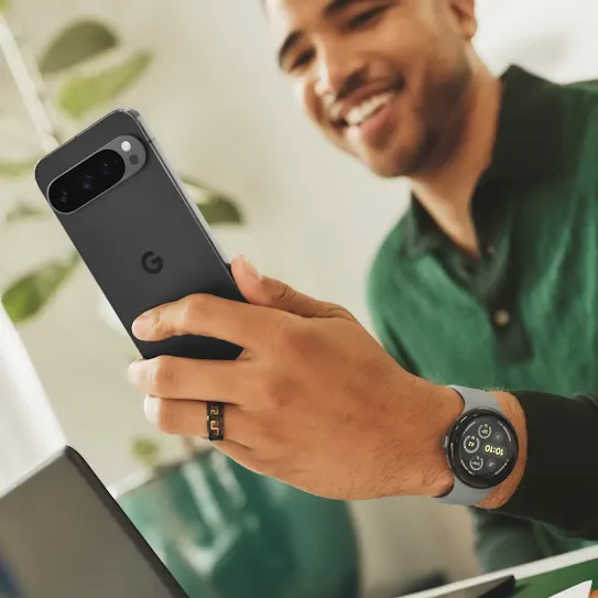 A young adult sitting at a desk smiles while looking at their Pixel 9 device.
