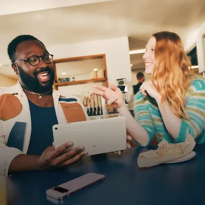 A man wearing glasses and a woman with long auburn hair are interacting in a cafe or casual workplace setting. The man is showing something on a tablet to the woman, who is smiling and looking at the man.