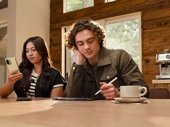 A woman is checking her Android phone. She is sitting next to a man who has a cup of coffee in front of him.