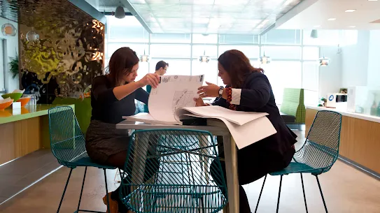 Two women sitting at a high-top table in a bright, modern office, focused on reviewing architectural floor plans together.