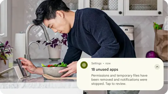 A person is preparing food by the kitchen sink while looking at their Android phone. A graphic overlay of a settings notification is placed on top of the image. It states that temporary files from unused apps have been removed and permissions have been reset.