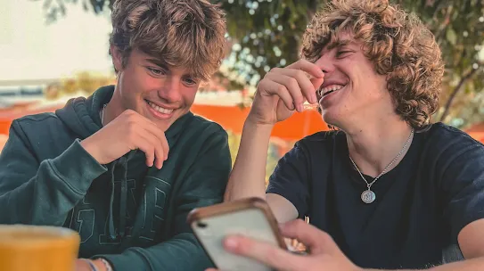 Two teenage boys with curly brown hair smiling and laughing together while looking at a smartphone at an outdoor cafe table.