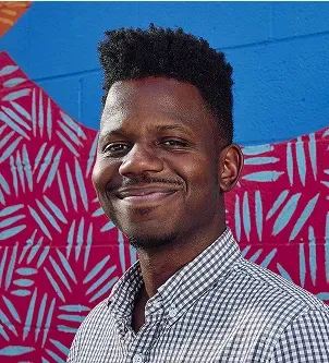 A portrait of Bernard Worthy, co-founder of LoanWell, smiling in a checkered shirt against a vibrant blue and pink patterned mural in North Carolina.