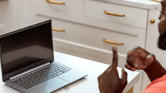 A Black man communicates in sign language to a person off camera at home. An open Chromebook is in front of him.